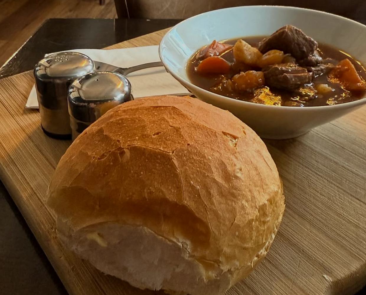 A bowl of beef stew with a fresh crusty roll on a wooden board